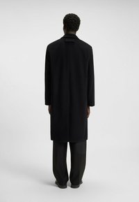 Black wool coat with a straight silhouette, long sleeves, and a back vent, worn over loose, dark trousers. Smooth fabric texture.