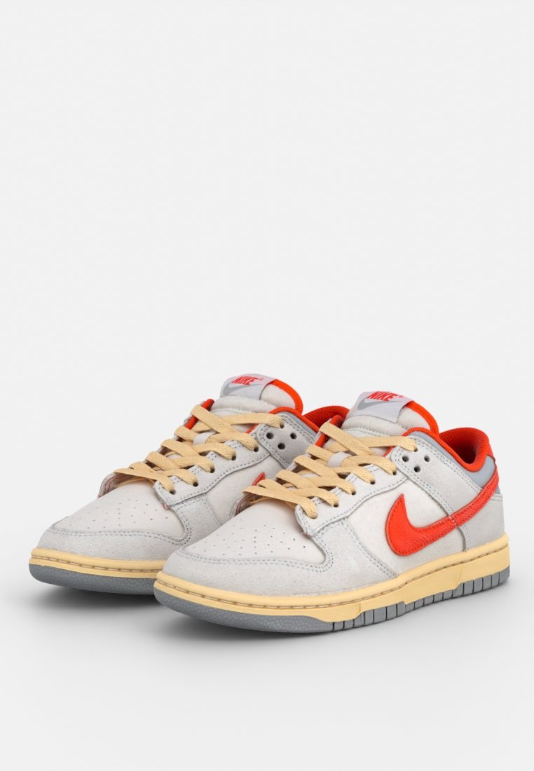 Le scarpe Nike presentano una tomaia in pelle grigia chiara, dettagli arancioni, lacci color crema e una suola intermedia gialla tessurizzata. Design della punta perforato.