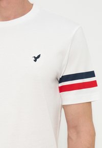 Λευκό κοντομάνικο μπλουζάκι με μικρό μαύρο λογότυπο κολιμπρί στο στήθος και ρίγες σε κόκκινο, λευκό και μπλε στη δεξιά μανίκι.