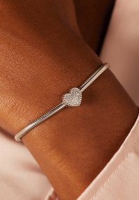 Zilveren armband met een gestructureerde band en een hartvormig middelpunt versierd met kleine, heldere stenen, gelegen op een bruine huidtint.
