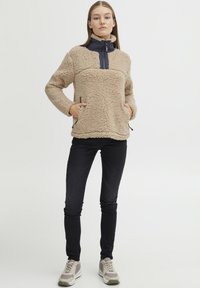 Pull-over en polaire beige avec col montant, patte de fermeture éclair marine et deux poches latérales. Porté avec un jean skinny noir et des baskets grises.