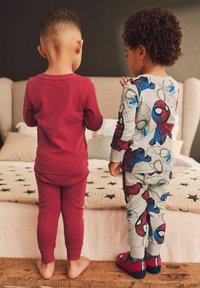 Zwei Kinder stehen mit dem Rücken zum Betrachter. Eines trägt ein einfarbig rotes Oberteil und passende Hose, während das andere einen grauen Schlafanzug mit Spider-Man-Motiven trägt.