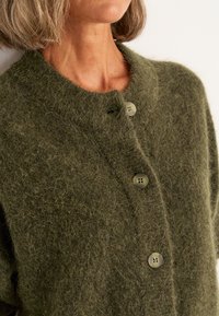 Olivgrüner Cardigan aus flauschiger Wolle, mit rundem Ausschnitt und drei Knöpfen mit subtiler glänzender Oberfläche, die eine weiche Textur präsentiert.