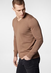 Brązowy sweter w serek z miękkiej dzianiny, dopasowany krój, ściągacze przy rękawach i u dołu, zestawiony z czarnymi dżinsami, tworzy swobodny wygląd.