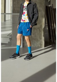 Zwarte ritsjack, wit en kleurrijk tie-dye T-shirt, blauwe geprinte shorts, blauwe sokken en zwarte sneakers met witte zolen.
