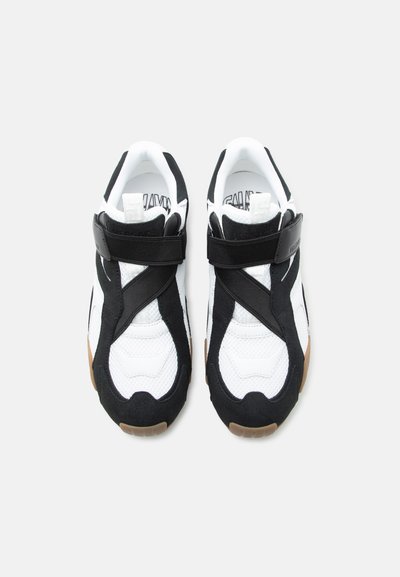 Ζευγάρι μαύρα και άσπρα αθλητικά παπούτσια με ρυθμιζόμενα λουράκια βέλκρο και σόλες χρώματος καουτσούκ, που φαίνεται από πάνω σε λευκό φόντο.