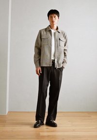 Chaqueta de cuadros gris con dos bolsillos en el pecho, llevada sobre una camiseta blanca, combinada con pantalones negros y zapatos negros. Textura suave, corte relajado.