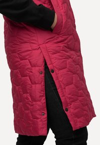 Gestepptes pinkes Weste mit sechseckigem Muster, seitlichen Schlitzen und Druckknopfverschlüssen, getragen über dunklen Jeans mit einem schwarzen Bündchen-Sweatshirt.