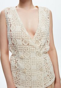 Gilet in pizzo off-white con scollatura a V profonda, motivi floreali e chiusura con bottoni in vita, caratterizzato da un design testurizzato e intricato.