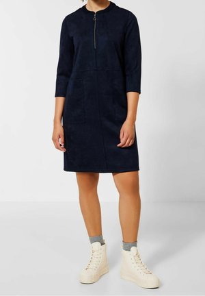 Žena v tmavě modrých šatech pod kolena s tříčtvrtečními rukávy, předním zipem, nášivkovými kapsami, šedými ponožkami a bílými kotníkovými teniskami.