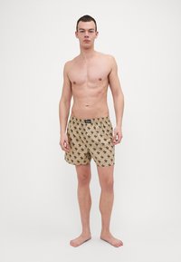 Beige boxerit dengan corak hitam berulang, pinggang elastik, dan potongan longgar, dibuat daripada fabrik lembut dan ringan.