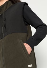 Fleece vest med en svart overdel og olivengrønn underdel. Har en glidelåslomme og et sideseam-etikett. Myk tekstur, uten ermer.
