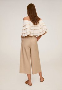 Vrouw met lang bruin haar staat met handen in de zakken, gekleed in een crèmekleurige off-shoulder top met lagen en ruffles, beige wijde, korte broek en bruine sandalen.