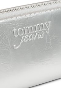 Ασημένιο πορτοφόλι χειρός με ένα ζαρωμένο υφή, ανάγλυφο λογότυπο "tommy jeans" και ασημένιο φερμουάρ κατά μήκος της κορυφής. Ομαλή επιφάνεια, ορθογώνιο σχήμα.