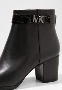 Schwarze Knöchelstiefel mit glänzender Lederoberfläche, quadratischem Absatz und einem Krokodillederstruktur-Gurt mit silberner MK-Logo-Akzentuierung.