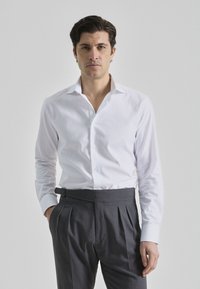 Camisa blanca de manga larga con cuello abotonado, combinada con pantalones grises oscuros con pliegues, que tienen bolsillos laterales y una cinturilla ajustada.