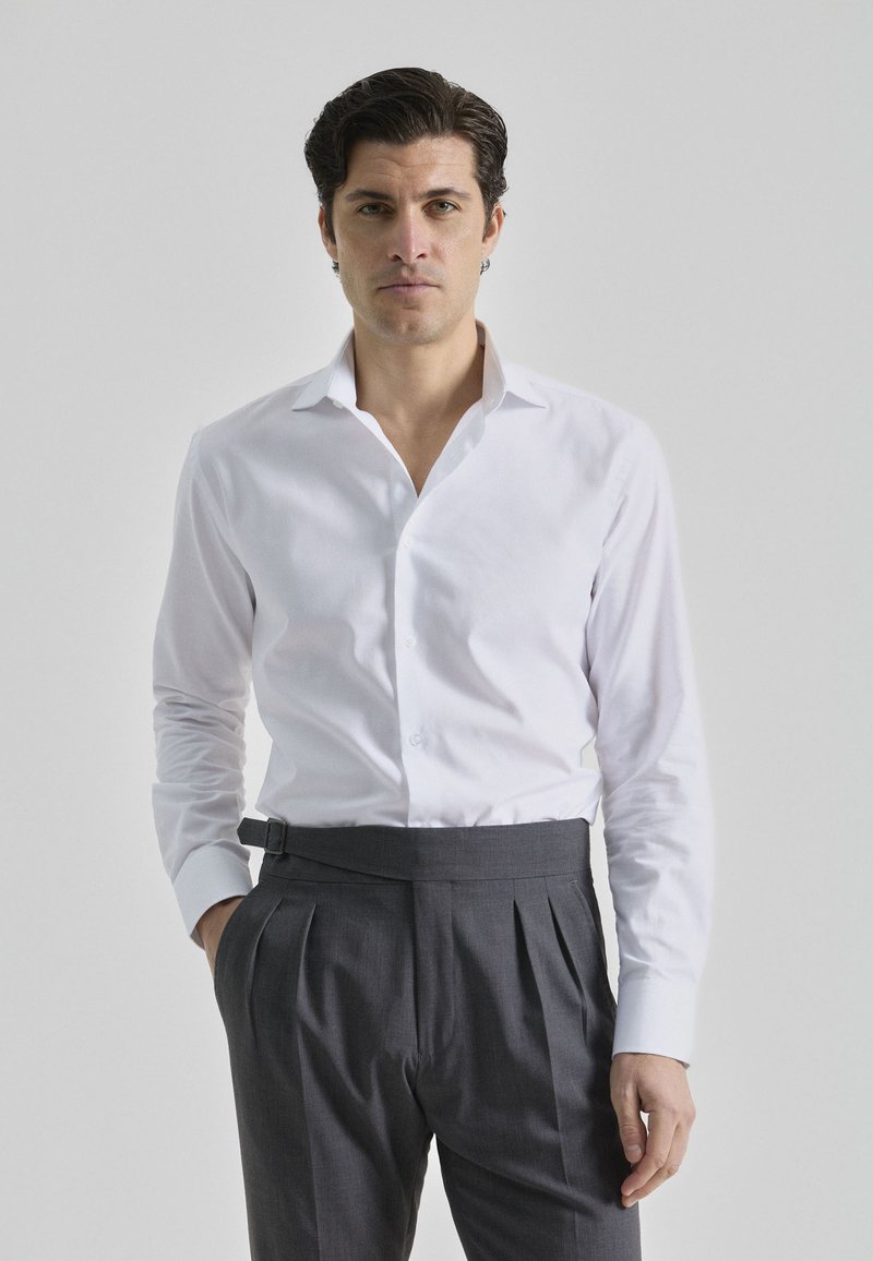 Camisa blanca de manga larga con cuello abotonado, combinada con pantalones grises oscuros con pliegues, que tienen bolsillos laterales y una cinturilla ajustada.