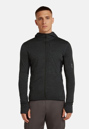 Mann mit kurzen Haaren, der eine dunkelgraue Zip-Down-Hoodie-Jacke und graue Hosen trägt, mit entspanntem Stand vor einem einfarbigen Hintergrund.