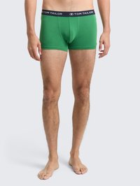 Uomo in piedi a piedi nudi, con indosso boxer verdi con vita nera etichettati "Tom Tailor", contro uno sfondo chiaro e semplice.