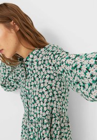 Il vestito floreale presenta uno sfondo verde con margherite bianche, una chiusura frontale con bottoni e maniche lunghe con texture arricciata.