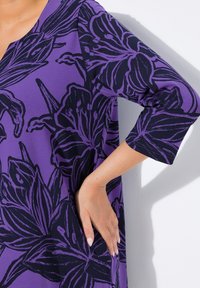 Die Hand einer Frau auf der Hüfte, trägt ein lila Kleid mit großen dunklen Blumenmuster und Dreiviertelärmeln vor einem weißen Hintergrund.