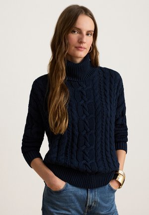 Femme aux longs cheveux bruns portant un pull à col roulé en maille torsadée bleu marine et un jean bleu, debout les mains dans les poches.