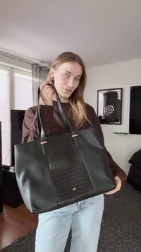 Jonge vrouw binnen met een grote zwarte tote bag met een gestructureerd middenpaneel, gekleed in een bruine trui en lichtblauwe jeans.