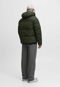 Dunkelgrüne Pufferjacke mit Kapuze und gestepptem Design. Kombiniert mit hellgrauen Hosen und Sneakers in neutralen Farbtönen.