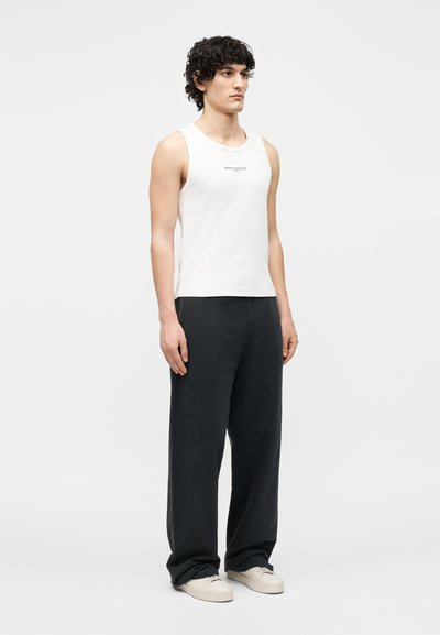 Νέος άνδρας όρθιος, φορώντας λευκό αμάνικο κορυφαίο με μικρό κείμενο και φαρδιά μαύρα παντελόνια με λευκά αθλητικά σε απλό φόντο.