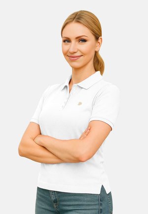 Weißes Polohemd mit Kragen, ausgestattet mit einer Knopfleiste mit drei Knöpfen und einem kleinen Logo. Trägt dazu eine blaue Jeans, die Arme verschränkt.