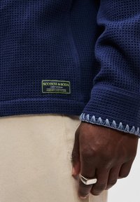 Σκούρο μπλε υφασμένο φανελάκι με ζικ ζακ μανίκια, που παρουσιάζει ετικέτα λογότυπου σε πράσινο χρώμα. Το χέρι φοράει ένα ασημένιο δαχτυλίδι και είναι ακουμπισμένο σε μπεζ ύφασμα.