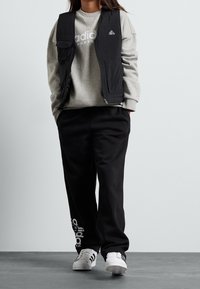 Černá vesta bez rukávů přes šedou mikinu s logem Adidas, doplněná černými tepláky a bílými botami s černými pruhy.