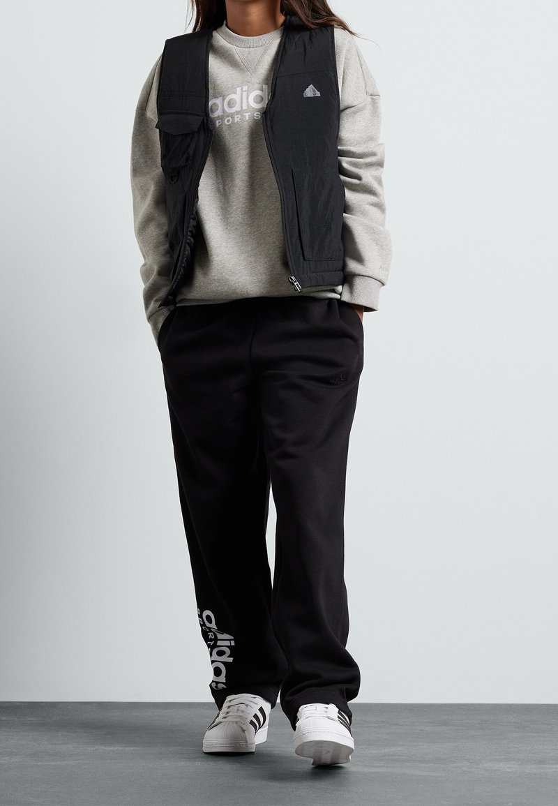 Černá vesta bez rukávů přes šedou mikinu s logem Adidas, doplněná černými tepláky a bílými botami s černými pruhy.