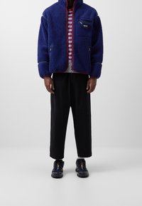 Blå fleecejakke med høj krave, lynlåslommer og røde detaljer. Båret over en grå- og rødstribet skjorte og parret med sorte bukser og sko.