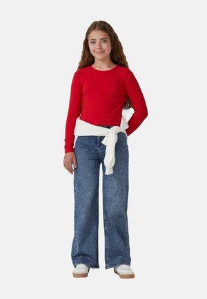 Top lungo a maniche lunghe rosso a coste abbinato a jeans azzurri a vita alta e gamba larga. Un maglione bianco è annodato in vita. Sneakers bianche completano l'outfit.