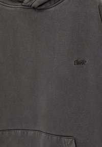 Sweat à capuche gris foncé montrant la partie avant supérieure avec un petit logo noir brodé et une partie de la poche kangourou en bas.