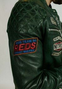 Veste de moto en cuir vert foncé avec texture matelassée aux épaules, ornée de patchs colorés : "ÉQUIPE REDS 84" et "NITRO", et fermetures éclair en argent.