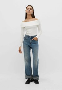 Weißes, schulterfreies Langarmoberteil mit geripptem Textur, kombiniert mit hochtaillierten, weit geschnittenen blauen Jeans mit umgeschlagenem Saum und Taschen.