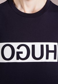 Marineblaue Baumwoll-T-Shirt mit einem weißen rechteckigen Streifen und großen schwarzen Buchstaben, die "HUGO" in der Mitte schreiben. Runder Ausschnitt.