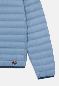 Svijetloplavi puffer jakna s kvadratnim dizajnom, glatkom teksturom i okruglom logotip oznakom na rubu u tamnoj boji.