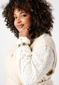 Crème-kleurige textuurvest met een kanten top met lange mouwen met intricate patronen; gouden armbanden accentueren de pols.