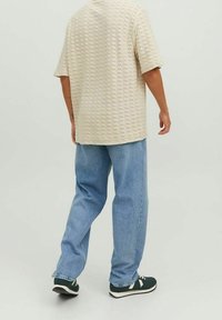 Persoon in een beige gestructureerd shirt met korte mouwen, lichtblauwe jeans en zwarte sneakers met witte zolen, staande op een effen achtergrond.