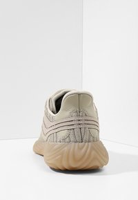 Beige sneakers med en texturerad stickad ovandel, böjd mellanfotssula och gummiyttersula. Har två dekorativa linjer tvärs över baksidan.
