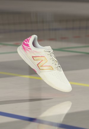 Weißer Sportschuh mit pinkfarbenen Akzenten und goldenem "N"-Logo, der über einem Hallensportfeld mit farbigen Linien schwebt.