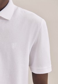 Polo bianca con tessuto a trama, maniche corte e un sottile logo ricamato sul lato sinistro del petto. Design classico del colletto.