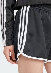 Närbild av en person i svarta träningsshorts och en långärmad topp med vita sidostråk, med handen avslappnad vid låret.