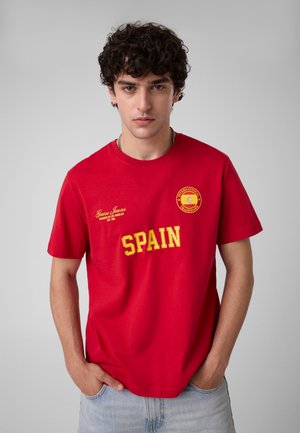 Giovane uomo con capelli ricci che indossa una maglietta rossa con scritta gialla "SPAIN" e jeans azzurri, in piedi davanti a uno sfondo grigio uniforme.