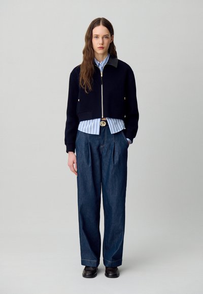 Chaqueta corta de marina con cremallera y detalles dorados, combinada con una camisa a rayas azules y pantalones de mezclilla holgados. Unos zapatos negros completan el conjunto.