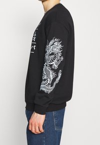 Felpa nera con un dettagliato disegno di un drago bianco sulla manica sinistra. Il tessuto è morbido, con polsini e orlo a costine.