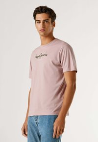 Roza bombažna majica s kratkimi rokavi in okroglim izrezom, na katerem je napisan tekst "Pepe Jeans London" v zeleni barvi na prsih. Obuta s modrimi kavbojkami.